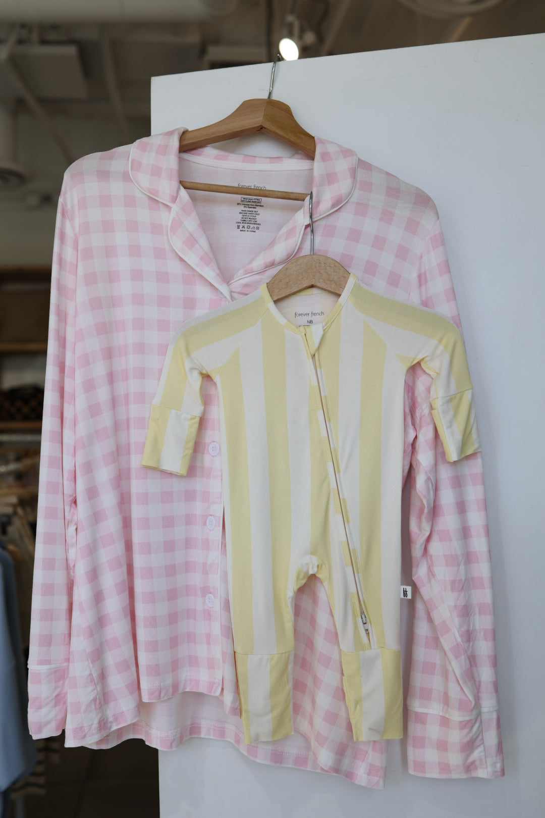 A Bamboo Zip Pajamas | Butter by forever french baby hangs on a wooden hanger next to a yellow and white striped breathable baby onesie, both displayed on a white wall in a store.