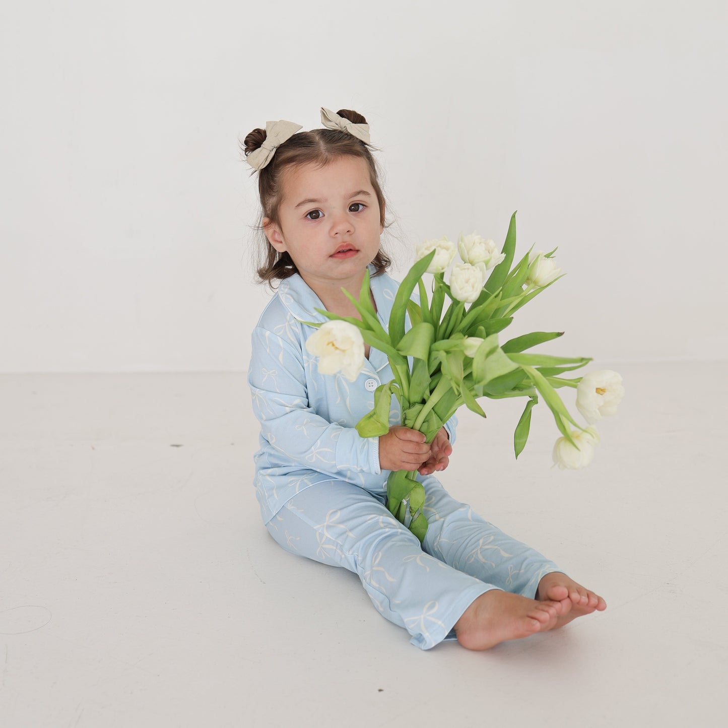 A young girl sits barefoot on a white background, wearing the forever french baby Girl’s Bamboo Button Up Lounge Set | Blue Bows and a white bow headband, holding white tulips. Hypo-allergenic comfort for toddlers included.