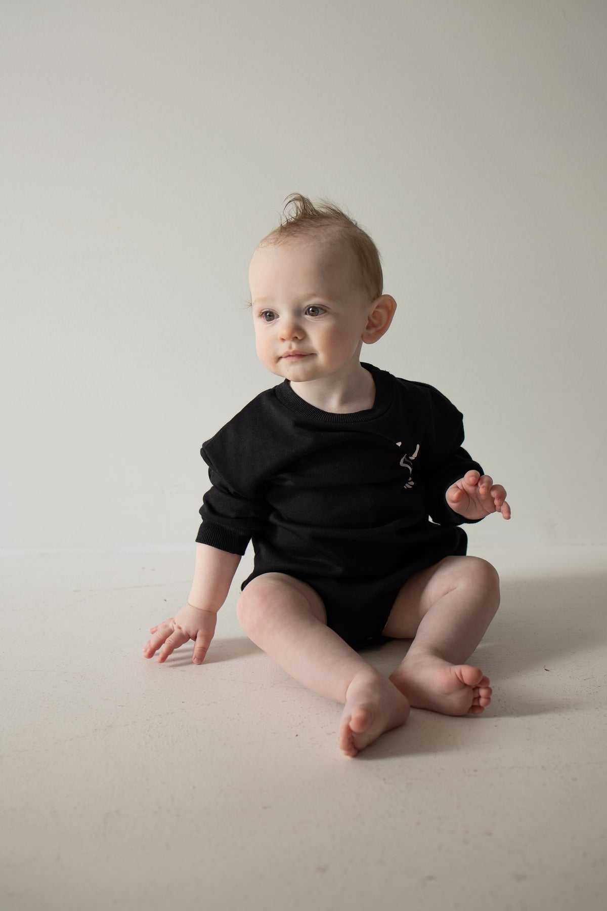 A baby with light skin and short hair sits on the floor wearing the Midnight Skelly Sweatshirt Romper from forever french baby, looking slightly to the left against a plain light-colored background.