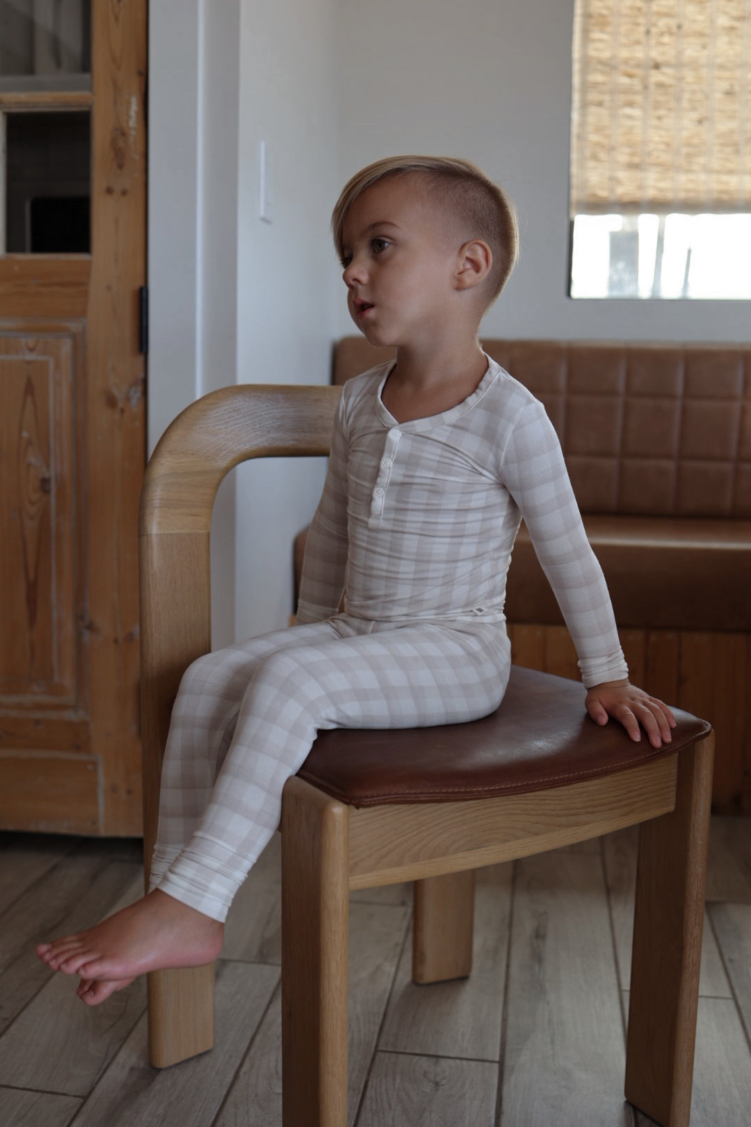 A young child with short, side-swept blonde hair sits sideways on a wooden chair, wearing forever french baby’s Bamboo Two Piece Pajamas | Gingham. These hypo-allergenic pajamas are perfect for sensitive skin in the cozy, wood-accented room.