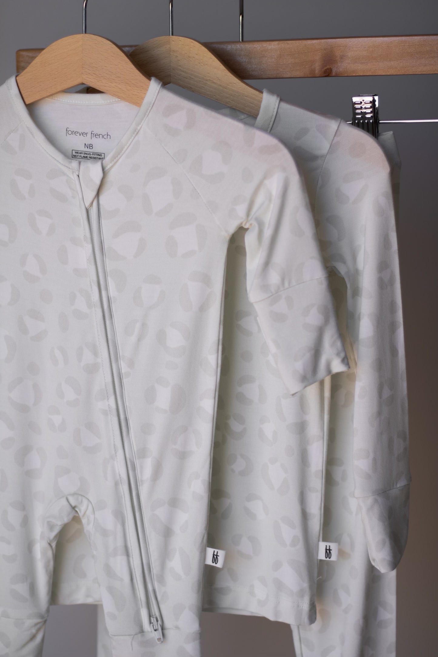 Two pale, patterned Bamboo Zip Pajamas | Meow by forever french baby hang on wooden hangers. Made from breathable fabric, these onesies feature subtle abstract prints and front zippers for comfort and style.