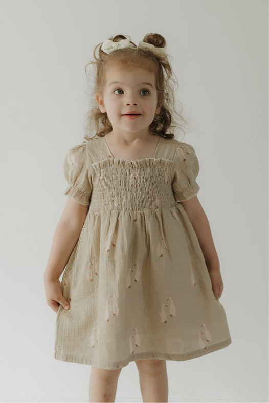 A young child with light brown hair in pigtails smiles off-camera, modeling the forever french baby Toddler Muslin Dress | Silly Goose—featuring breathable cotton muslin, short puffed sleeves, and subtle patterns—against a plain white background.