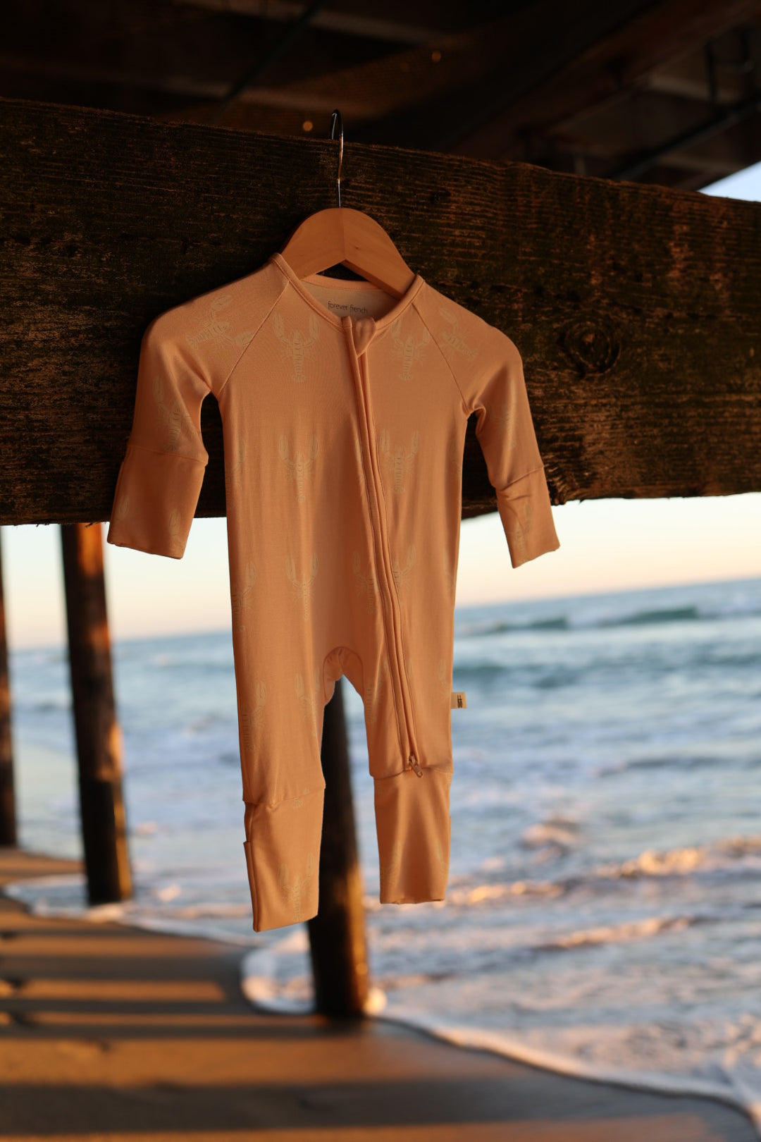 The forever french baby Bamboo Zip Pajamas | Lobster, a hypoallergenic long-sleeve and long-leg onesie in light pink, hangs on a wooden hanger beneath a pier with sunset waves and sandy beach in the background.