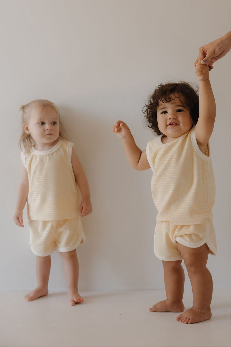 Two toddlers wear matching Forever French Baby Terry Tank Short Set in Creamsicle, standing barefoot against a plain wall—one holds an adult's hand, while the other stands with hands at her sides.