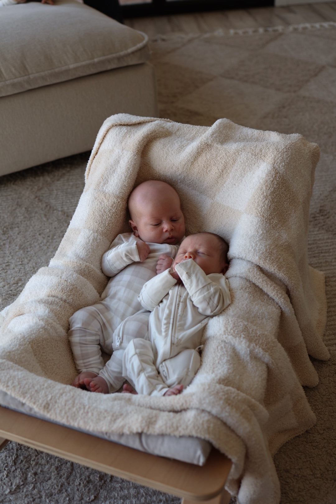 Two infants in forever french baby Bamboo Zip Pajamas | Gingham lie side by side on a padded seat, covered with a beige blanket. They appear to be sleeping or resting, with a soft rug and couch in the background.