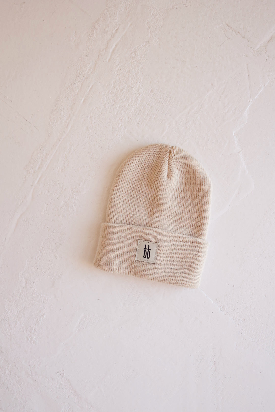 An oatmeal-toned ff Knit Beanie with a folded brim and ribbed design rests on a textured white surface. The small rectangular tag on the brim features Forever French Baby's minimalist, stylized "bb" logo in black.