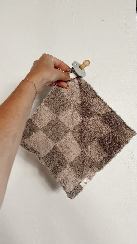 A hand holds the Lovey | Wavy Checker from Forever French Baby, a whimsical cloth with a pacifier attached. The soft microfiber feather yarn elegantly drapes against a white background, capturing the essence of our Forever French Baby collection.
