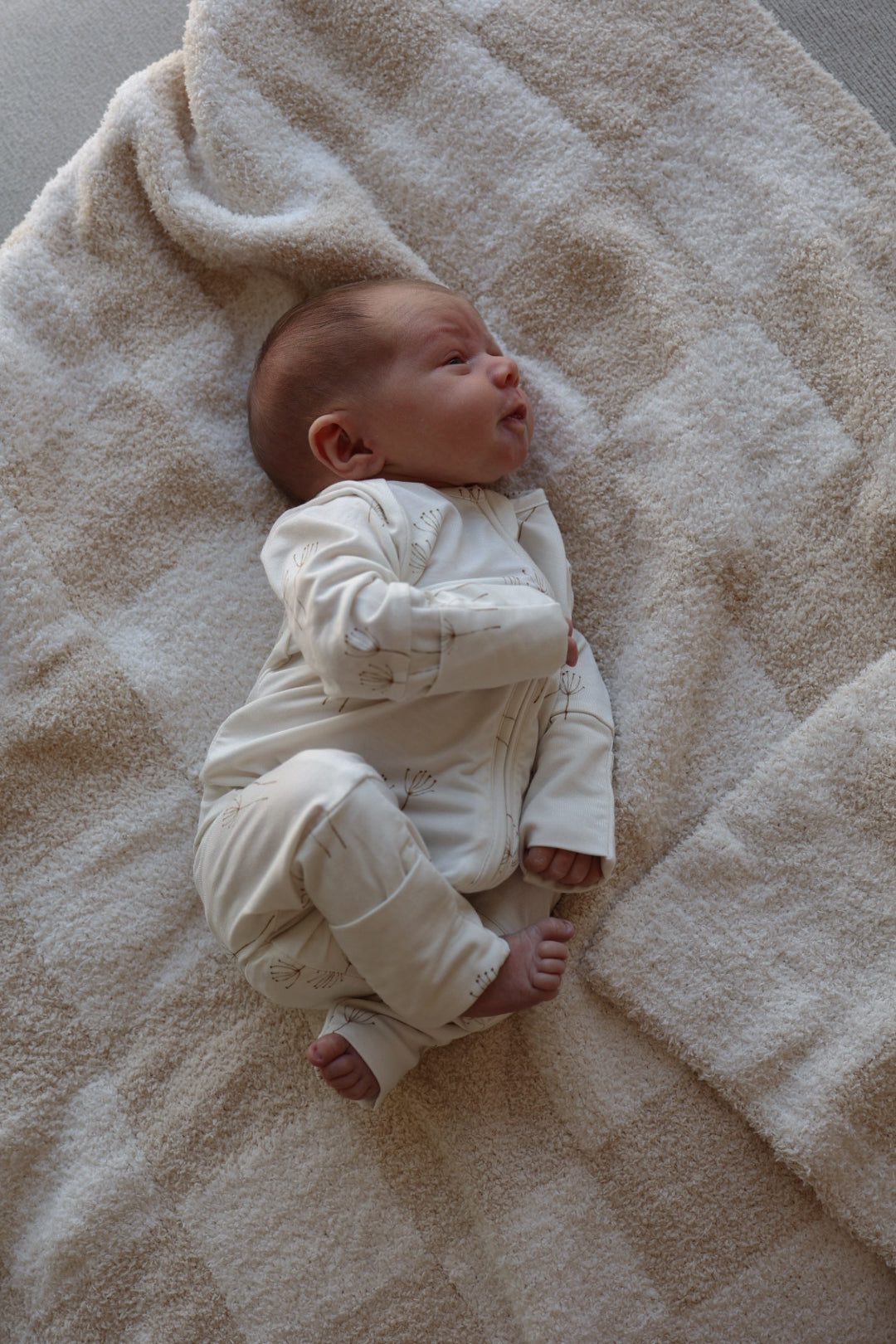 A baby in forever french baby's Bamboo Zip Pajamas | Dandelion lies on a soft, beige textured blanket, gazing upward with one arm bent and tiny feet peeking out.