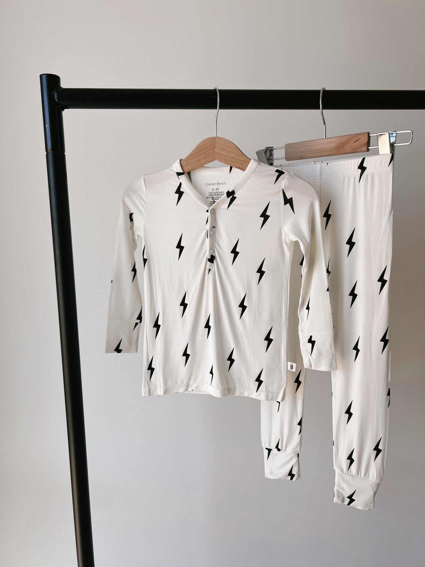 A chic set of Bamboo Two Piece Pajamas | Black & White Lightning Bolt from forever french baby is displayed on a wooden hanger, suspended from a sleek black metal clothing rack. This adorable children's pajama set, featuring black lightning bolt patterns on a white fabric, includes a long-sleeve button-up top and matching pants. Made from hypo-allergenic bamboo material, it stands out against the plain white wall background.