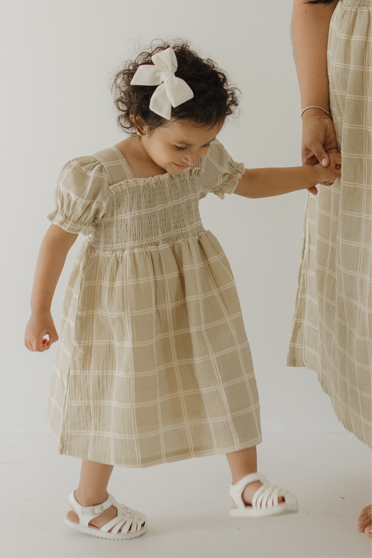 A young girl in the “Toddler Muslin Dress | Linen Grid” by forever french baby holds an adult’s hand. Both wear matching lightweight muslin dresses against a plain light background.