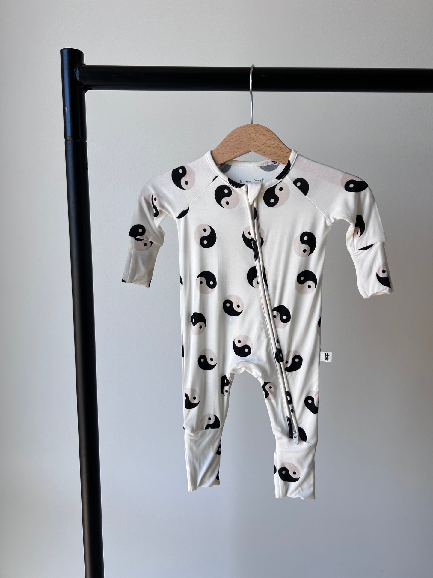 A bamboo zip onesie with long sleeves, showcasing a black and white yin-yang symbol pattern, hangs on a wooden hanger suspended from a black metal clothing rack against a plain white background. This piece is part of our Baby and Toddler clothing collection by forever french baby, known as the Bamboo Zip Pajamas | Black & White Yin Yang.
