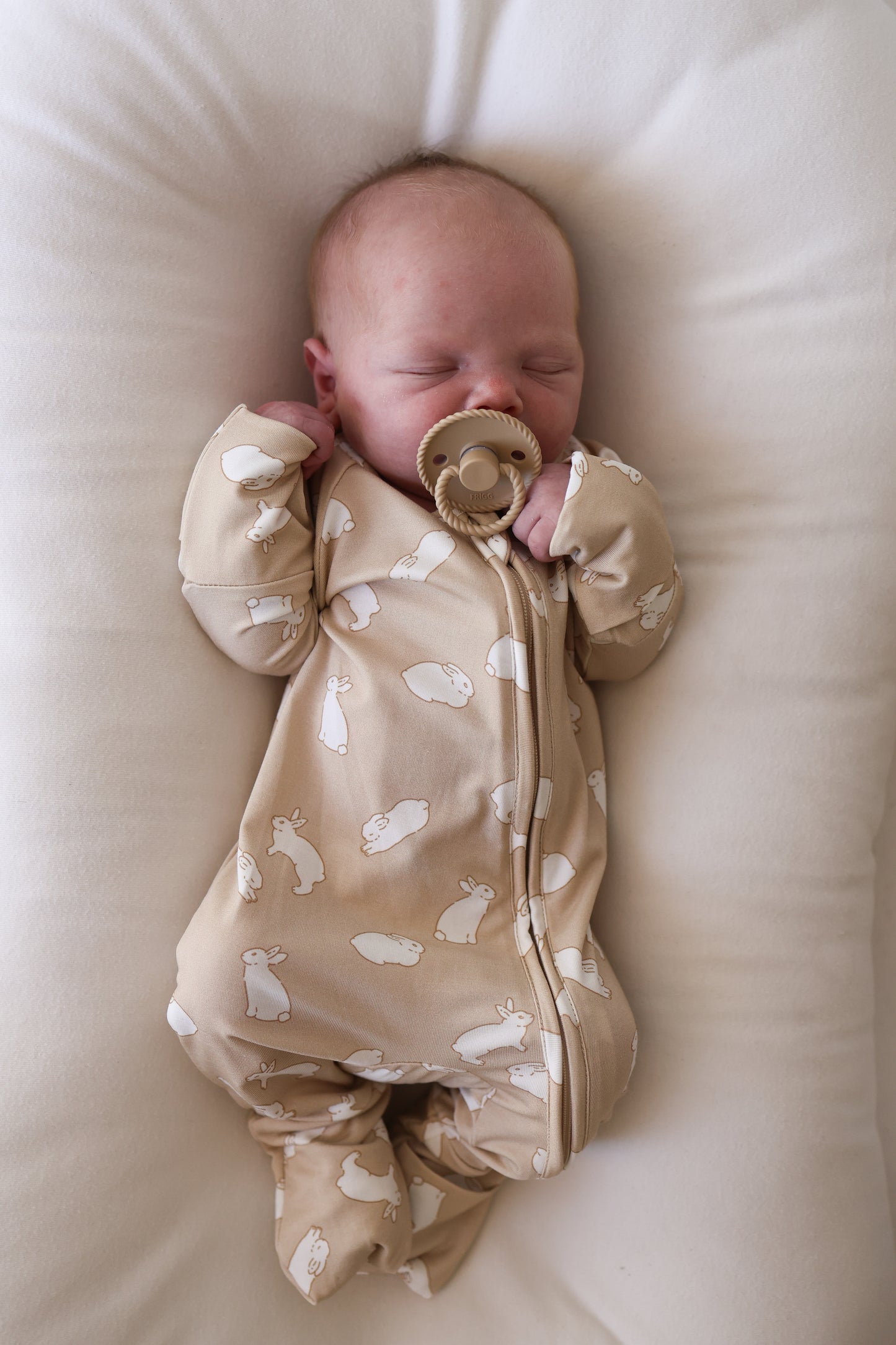 A newborn sleeps peacefully on a white cushion, wearing forever french baby's Bamboo Zip Pajamas | Bunnies—a cozy beige onesie with bunny prints—and using a tan pacifier, hands near its face and eyes closed.