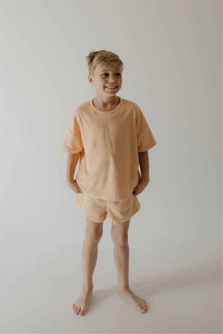 A young blond boy stands barefoot against a white background, smiling and wearing the forever french baby Child Terry Short Set in Sherbert, his hands in his pockets—a perfect look for family moments.