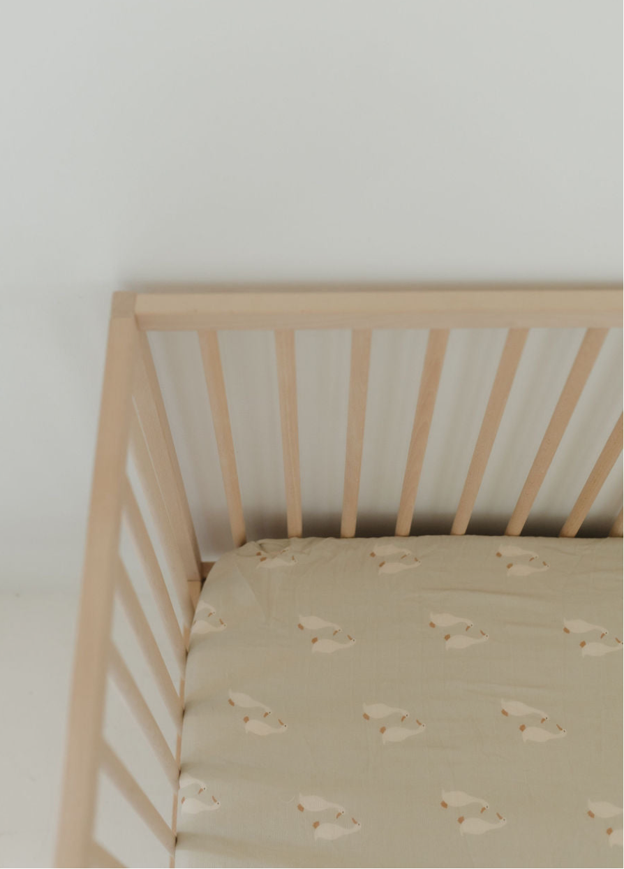 A wooden baby crib with light-colored slats, fitted with the forever french baby Muslin Crib Sheet | Silly Goose, featuring a simple sheep pattern on a beige background.