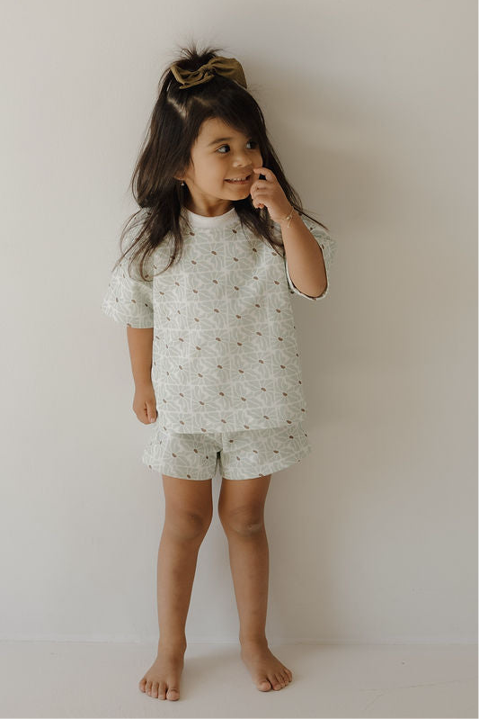 A young girl with long dark hair and a large bow stands barefoot, smiling as she touches her nose. She wears the forever french baby French Terry Short Set | Coast, featuring a soft short sleeve top and matching shorts with a subtle FF logo pattern.