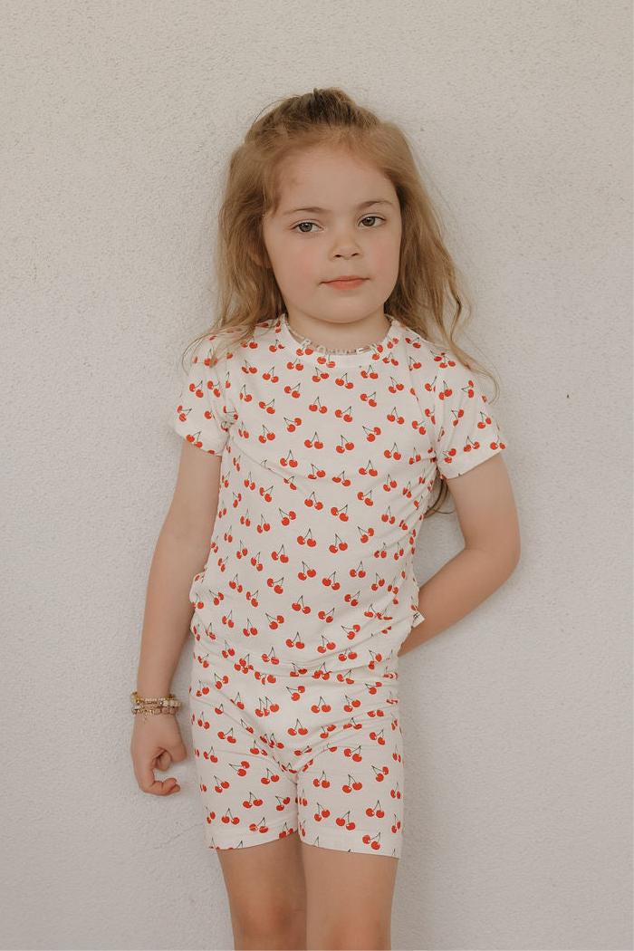 A young girl with long light brown hair stands against a white wall, wearing the forever french baby Bamboo Short Set | Sweetheart Cherries—cream-colored pajamas with a red cherry print. She has a neutral expression and her left hand on her hip.