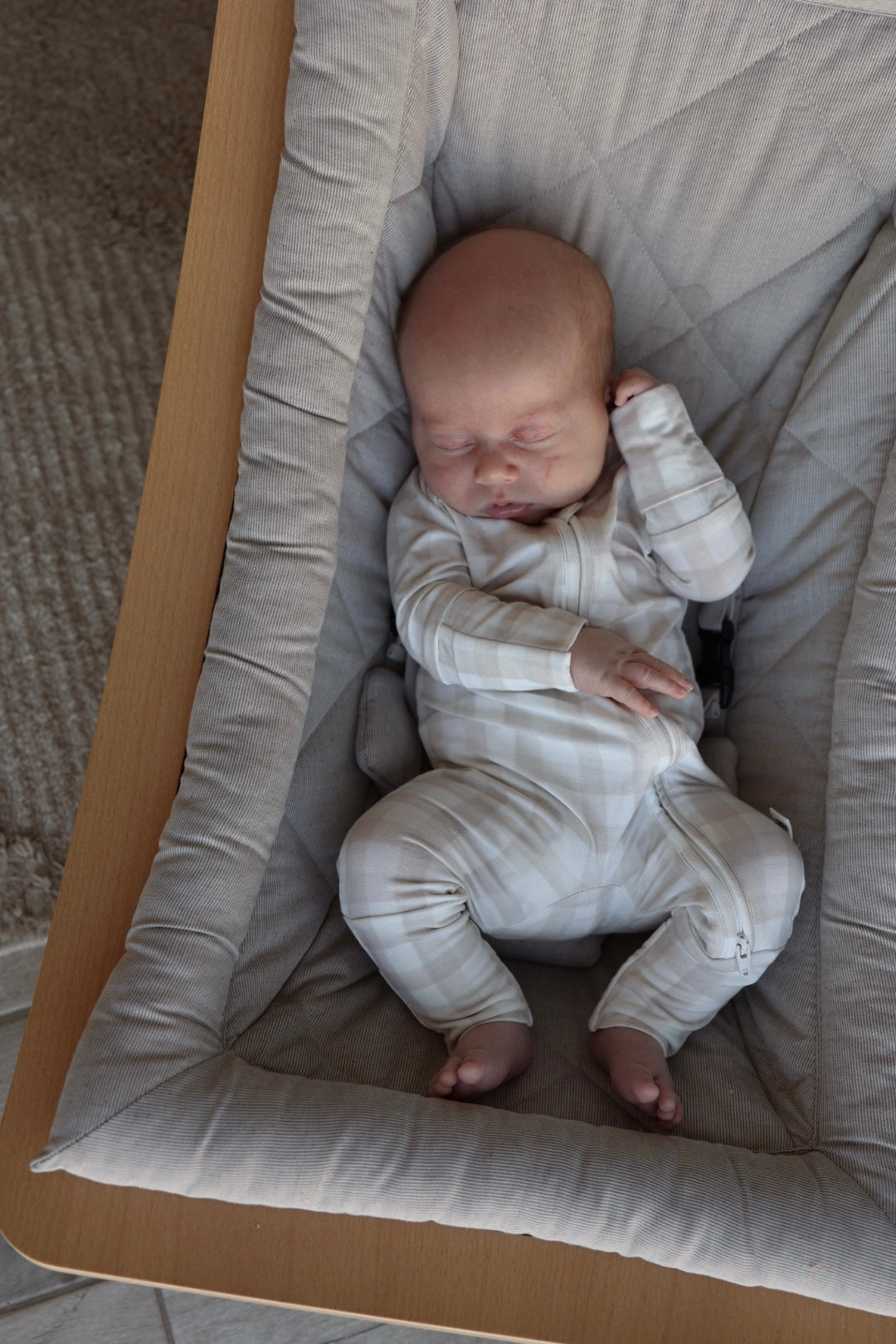 A newborn sleeps peacefully in a cushioned baby lounger, dressed in forever french baby Bamboo Zip Pajamas | Gingham—breathable and hypoallergenic pajamas with a white and beige checkered design.