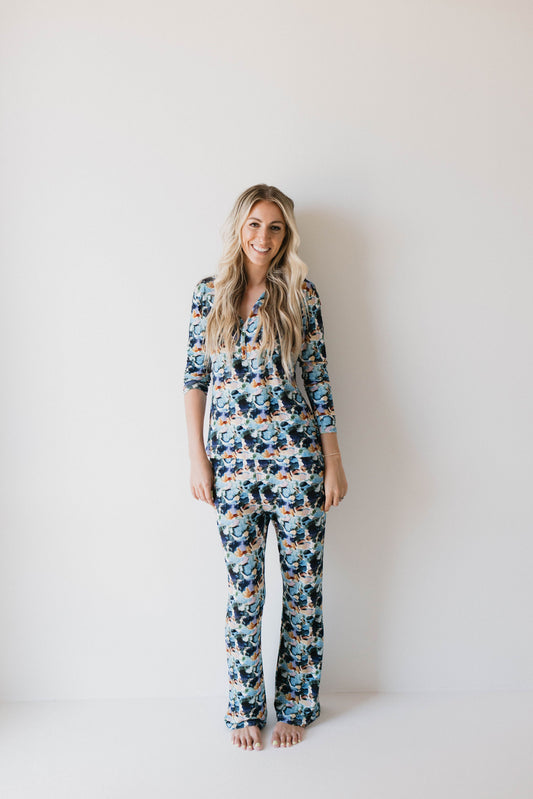 A woman with long blonde hair stands barefoot against a plain white wall, wearing the Women's Bamboo Pajamas in Charli Print from forever french baby. She is smiling, her hands relaxed by her sides, radiating calm and perhaps even promoting autism awareness through her serene presence.