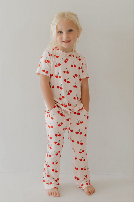 A young child with long blonde hair stands barefoot in the forever french baby Toddler Short Sleeve Bamboo Lounge Set | Cherry Orchard, featuring a white bamboo fabric with a red cherry pattern, hands in pockets, enjoying its breathable comfort.
