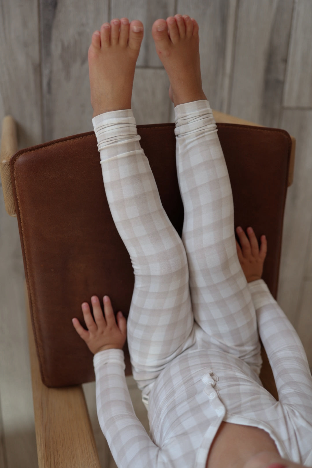A young child wearing forever french baby Bamboo Two Piece Pajamas | Gingham in cream and white lies on a chair with a brown cushioned seat and wooden armrests, legs raised and feet resting on the chair's backrest.