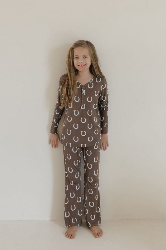 A young girl with long light brown hair smiles barefoot against a plain wall, wearing forever french baby’s Youth Flare Bamboo Pajamas | Giddy Up—brown pajamas with white horseshoe print, breathable fabric, long sleeves, and loose pants.