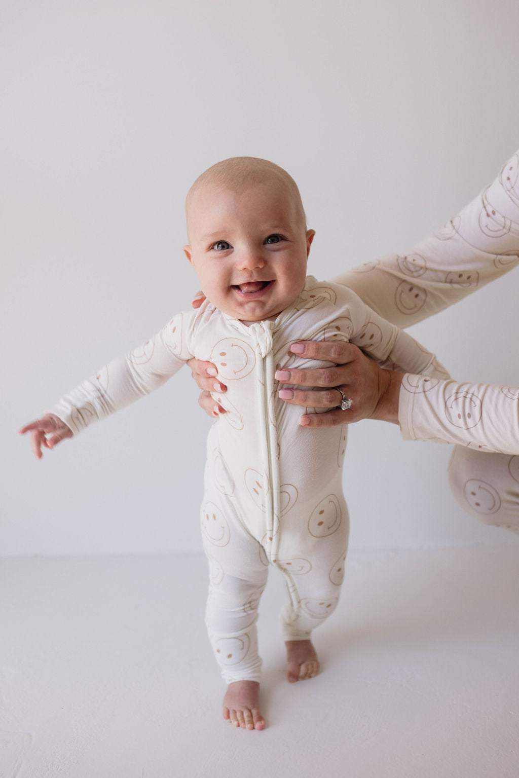 A smiling baby stands with support from an adult's hands. The baby is wearing Forever French Baby's Bamboo Zip Pajamas in the Just Smile pattern, which features a premium cream-colored fabric adorned with smiling faces. The plain white background highlights the baby's cheerful expression.
