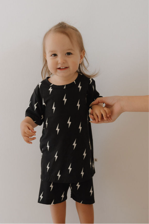 A toddler wearing the forever french baby Bamboo Short Set in Midnight Lightning Bolt—featuring a black shirt and shorts with white lightning bolts—stands against a plain background, smiling and holding an adult’s hand.