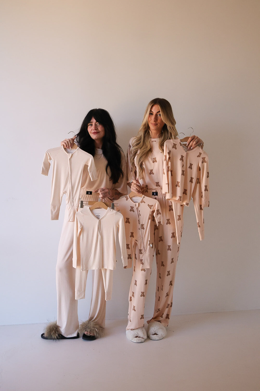 Two women in pajamas and slippers stand against a plain wall, each holding up matching forever french baby Bamboo Two Piece Pajamas | Sadie Stripe—light, neutral-toned bamboo sleepwear that’s gentle on sensitive skin and perfect for cozy nights.