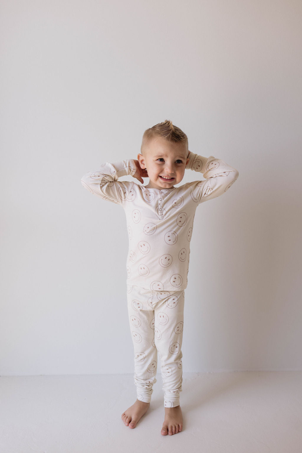 A young child with short, light-colored hair stands barefoot against a plain white background. The child is smiling and wearing breathable Bamboo Two Piece Pajamas from forever french baby, adorned with a pattern of smiley faces. Their hands are placed behind their head.