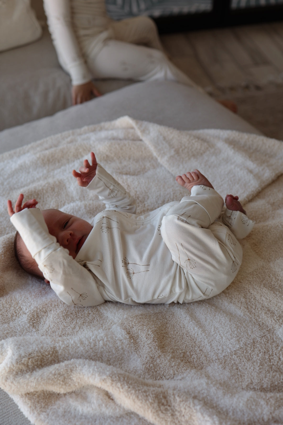 A baby in forever french baby Bamboo Zip Pajamas | Dandelion lies on a soft cream blanket with arms raised, while another child in matching pajamas sits partially visible on a couch in the background.