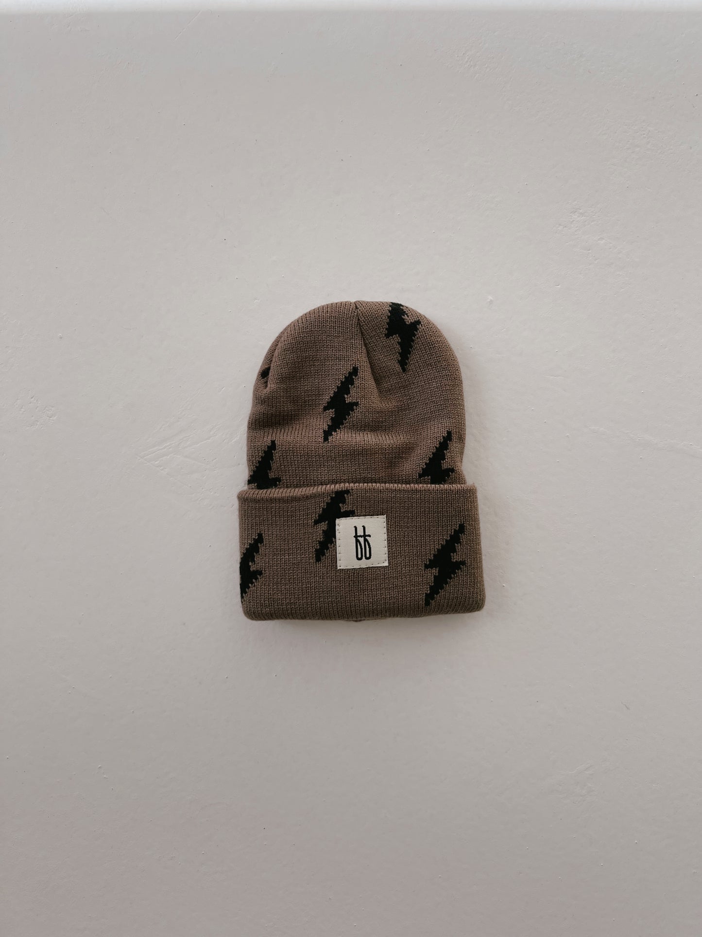 Against a plain white background, the ff Knit Beanie from forever french baby is prominently displayed. This brown beanie features black lightning bolt patterns and a classic ribbed design. It is adorned with a small rectangular tag bearing the Forever French Baby logo sewn onto the front cuff.