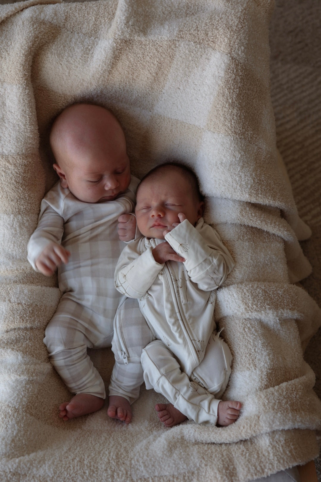 Two babies lie side by side on a beige and cream plaid blanket, wearing forever french baby Bamboo Zip Pajamas | Gingham; one baby is awake with open eyes, while the other sleeps peacefully with hands near its face.