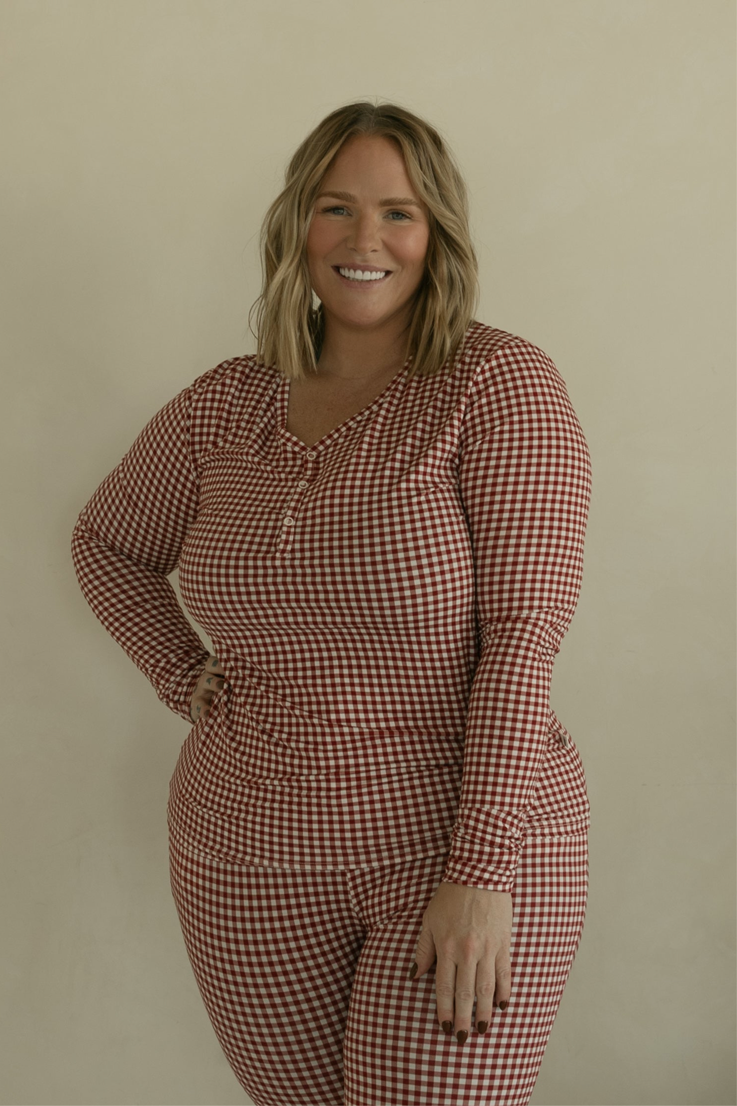 A smiling woman with blonde, shoulder-length hair wears forever french baby's Women's Bamboo Pajamas in Red Gingham, standing against a plain beige background.