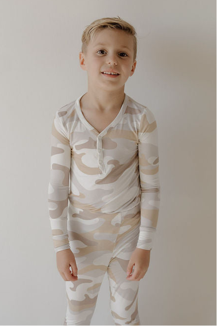 A young boy with short blonde hair smiles while standing against a plain wall, wearing forever french baby's Bamboo Two Piece Pajamas | Camo 2.0—a comfy, breathable beige and white camo-patterned sleepwear set.