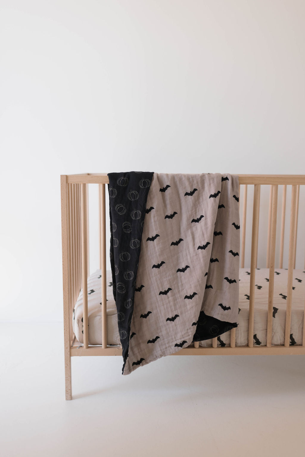 A wooden baby crib against a white background, featuring nursery bedding including a beige mattress adorned with black bat print sheets. Draped over the side of the crib is a Muslin Quilt from forever french baby—one side showcasing a black and white circular pattern, while the other side matches the bat print of the sheets.