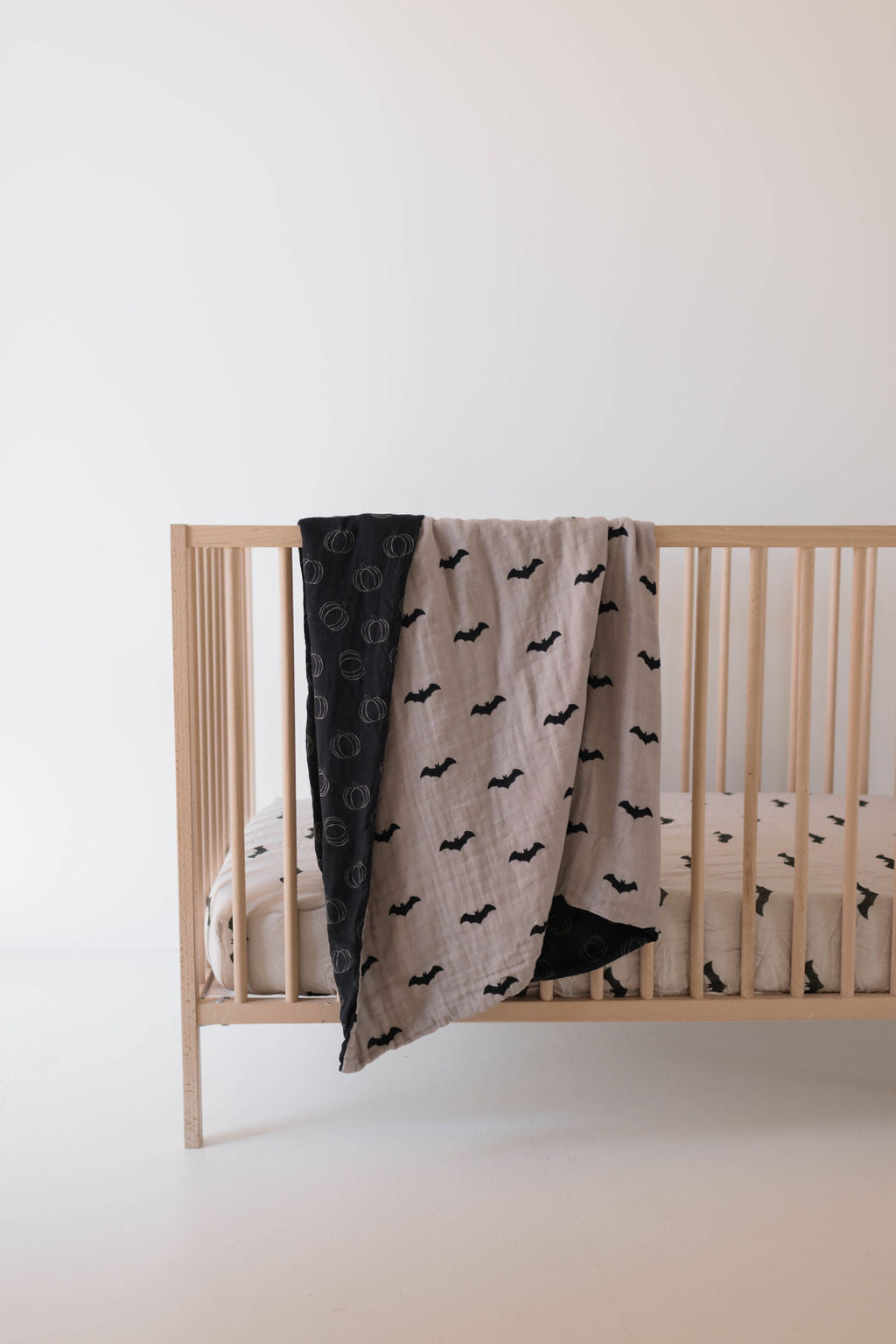 A minimalistic nursery setup with a light wooden baby crib. The crib contains the “Muslin Quilt | It's Bats!” by forever french baby, featuring a pattern of black bats on muslin cotton. A similar quilt is draped over the rail. The background is a plain off-white wall, contributing to the room's simplicity.