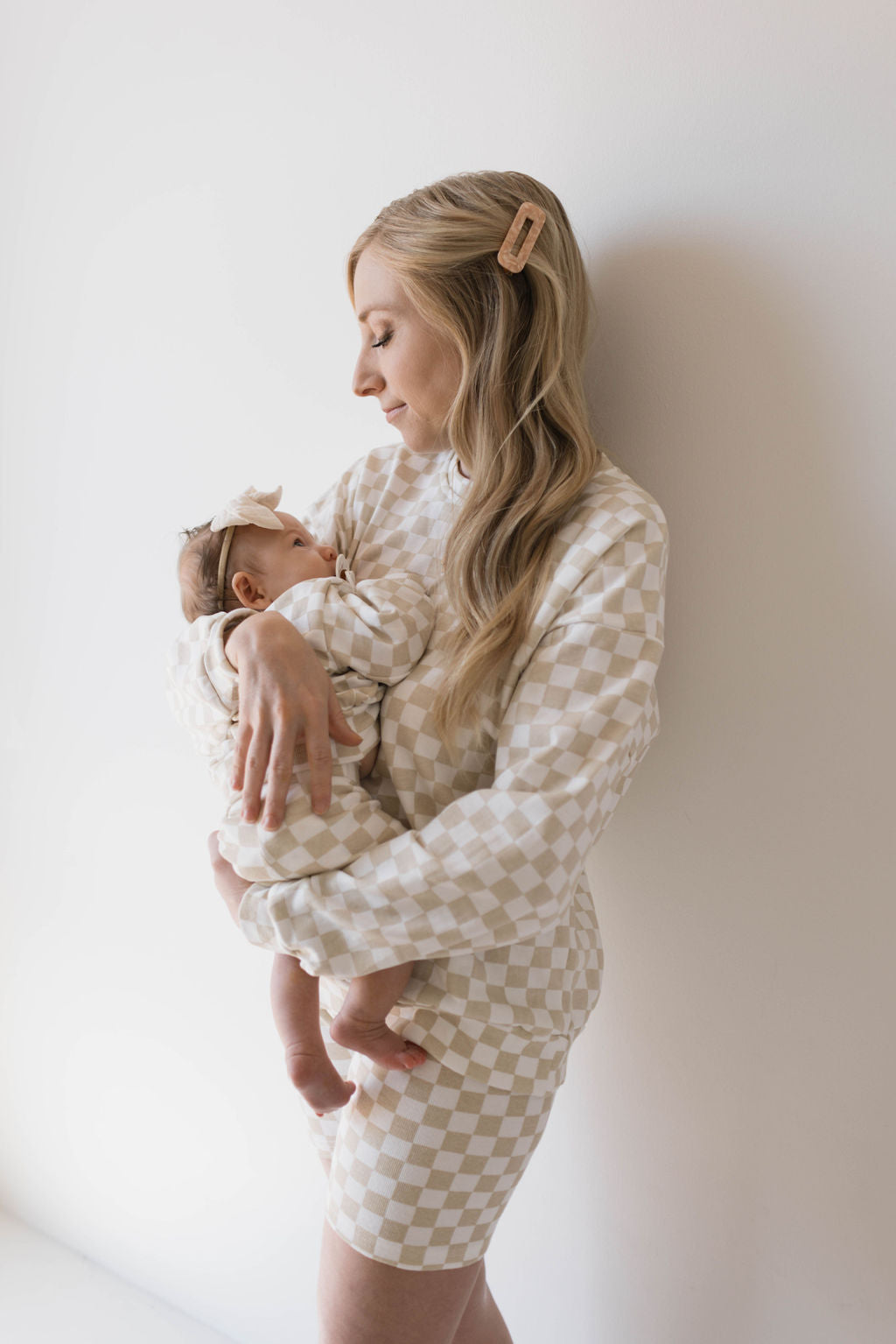 A woman with long blonde hair, secured with a hair clip, lovingly cradles a baby. Both are wearing matching Chai Checkerboard mama-and-me outfits from the brand forever french baby. The background is a plain, light-colored wall. The woman gazes softly at the baby, who is peacefully resting.