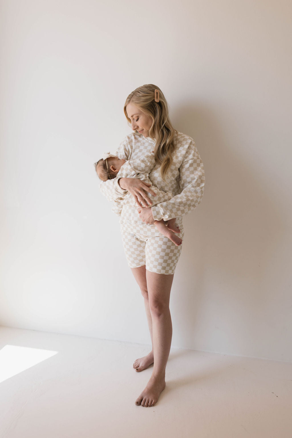 A woman with long blonde hair stands barefoot against a plain white wall, holding a baby. Both wear matching mama-and-me beige and white checkered outfits from forever french baby, including ribbed shorts. The woman gazes down at the baby, who is resting peacefully in her arms, wearing the Children's Long Sleeve Short Set in Chai Checkerboard.