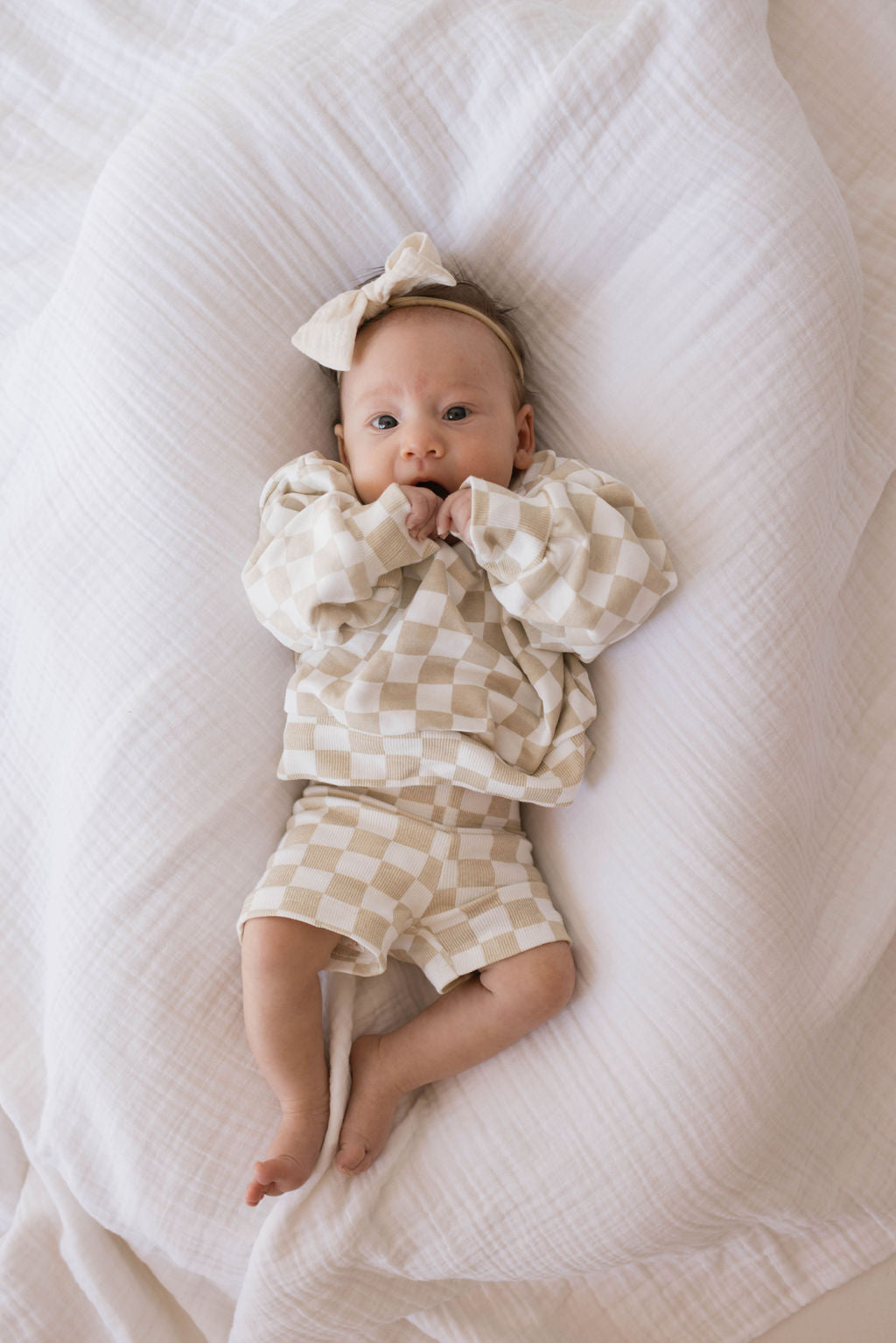 A baby lies on a soft white blanket, wearing the forever french baby Children's Long Sleeve Short Set in a charming Chai Checkerboard pattern, complete with a matching headband. The baby is looking up with hands near their mouth, making it perfect for those adorable mama-and-me moments.