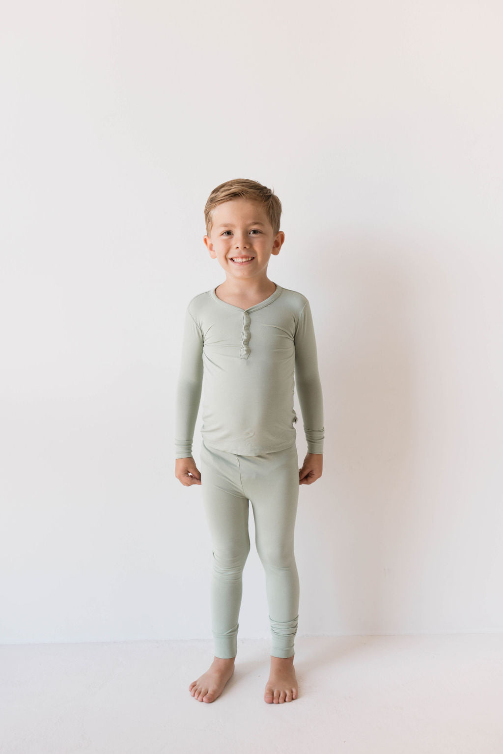 A young child stands barefoot against a plain white background, smiling at the camera. They are wearing the forever french baby Bamboo Two Piece Pajamas in Pistachio from ff club, featuring a light green, long-sleeved top and matching pants that ensure comfort and breathability.