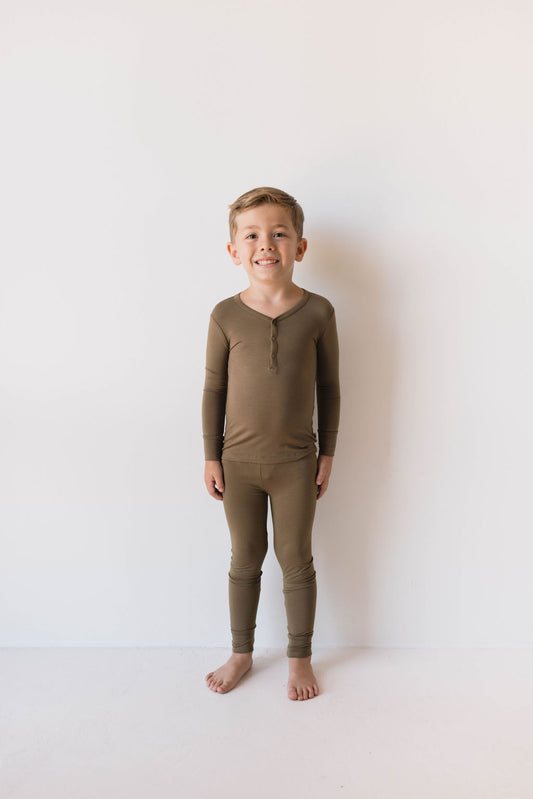 A young boy stands barefoot on a white surface against a plain white background, smiling in his Forever French Baby Bamboo Two Piece Pajamas from ff club. The matching long-sleeve shirt and pants are a solid olive green color and made from hypo-allergenic bamboo fabric. His hands are relaxed at his sides.