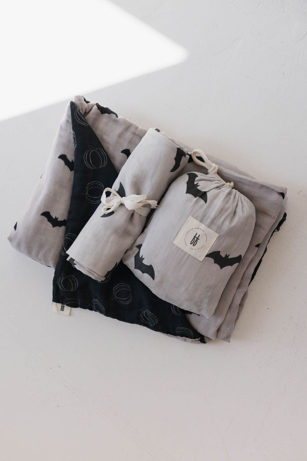 A bundle of neatly folded fabric items, including the "Muslin Quilt | It's Bats!" by forever french baby and two smaller cloth bundles in gray and black, all featuring bat and spiral designs. One of the muslin cotton cloth bundles has a white tag attached with the logo. The items are placed on a white surface.