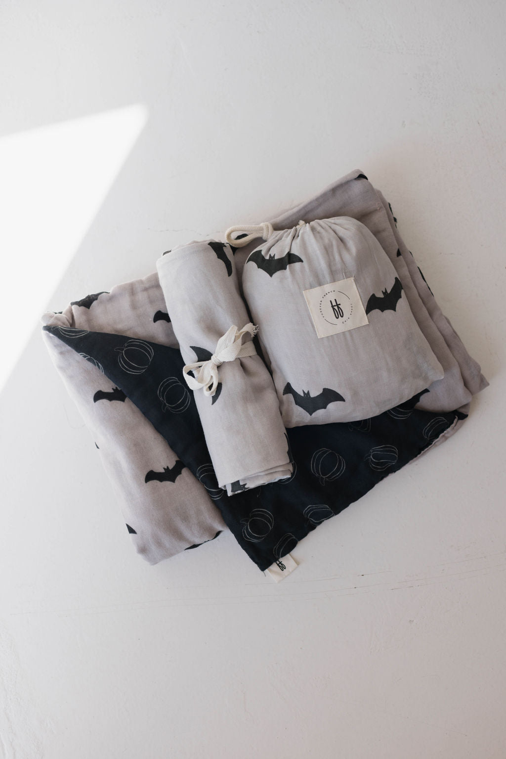 Displayed is a neatly folded set of soft, grey and black baby blankets from forever french baby. The "Muslin Quilt | It's Bats!" features one blanket with bat patterns and another adorned with spiral designs. A small, matching fabric bag with a drawstring rests atop the nursery bedding.