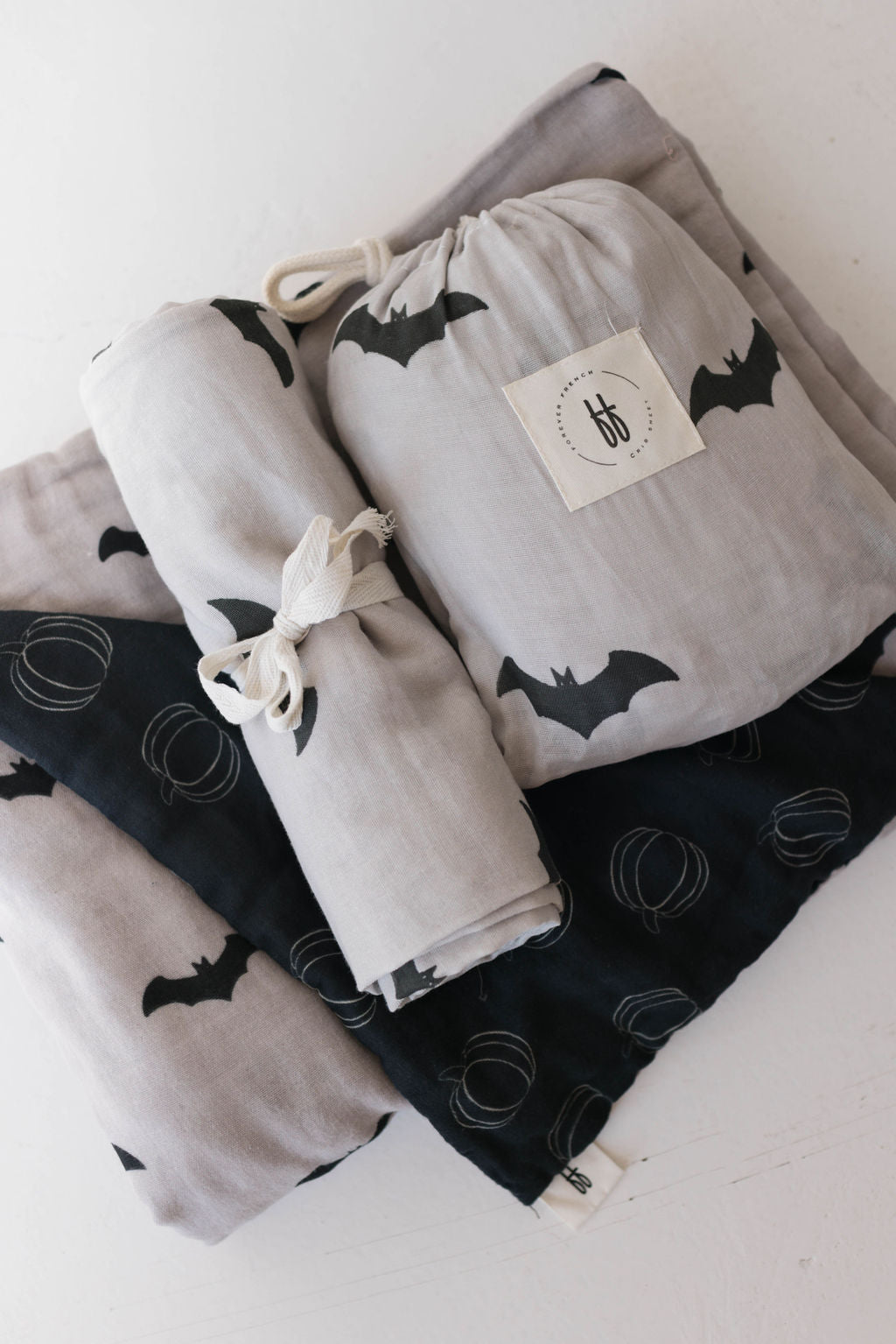 A collection of folded fabrics and cloths, showcasing Halloween-themed prints such as bats and pumpkins in gray and black hues. Ideal for creating a baby quilt or nursery bedding, one fabric roll from the "Muslin Quilt | It's Bats!" range by forever french baby is elegantly tied with a ribbon, while another is neatly packed in a drawstring bag with a printed label.