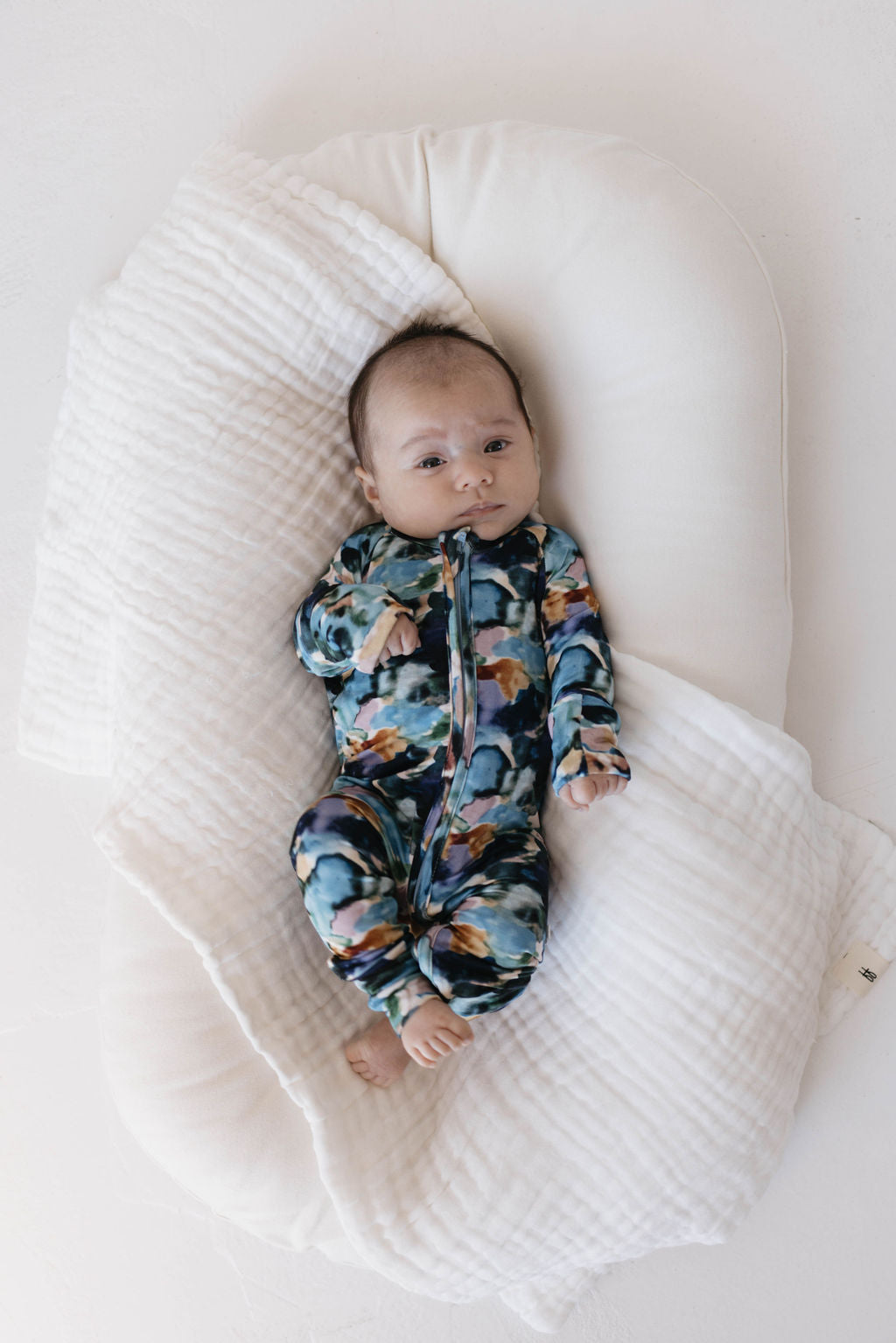 A baby in a cheerful Charli Print onesie from forever french baby lies on a soft, white cushion. Partially wrapped in a white, waffle-textured blanket, the baby looks upward with wide eyes, their bare feet resting on the cushion. The plain white background accentuates the vibrant Bamboo Zip Pajamas and cozy bamboo clothing.