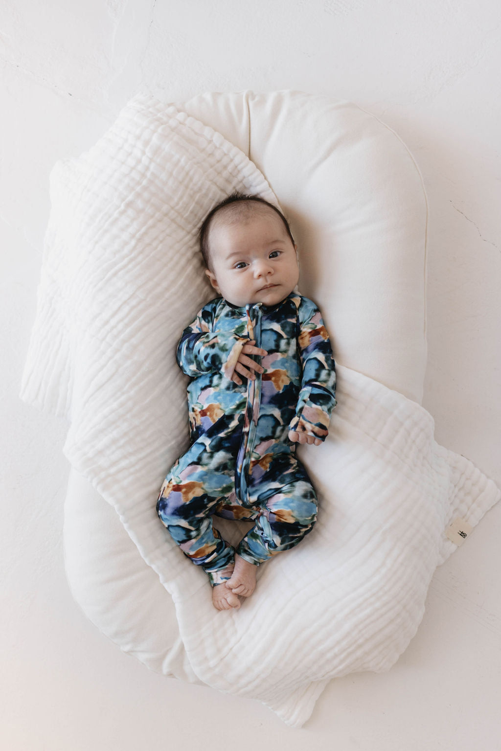 A baby rests peacefully on a white cushioned surface, swaddled in a white blanket. The baby is dressed in vibrant, floral-patterned Bamboo Zip Pajamas | Charli Print by forever french baby, made from soft bamboo fabric and gazes upward with a serene expression. The background is light and neutral.