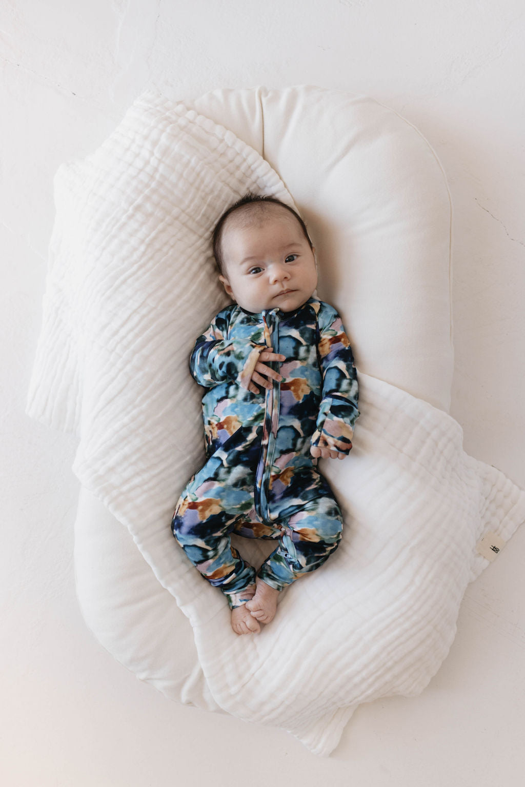 A baby lies on a soft, white blanket, looking up. The baby is wearing forever french baby's Bamboo Zip Pajamas in the Charli Print, featuring colorful patterns with blue, green, and brown hues. The baby's eyes are open and tiny toes peek out from the onesie.