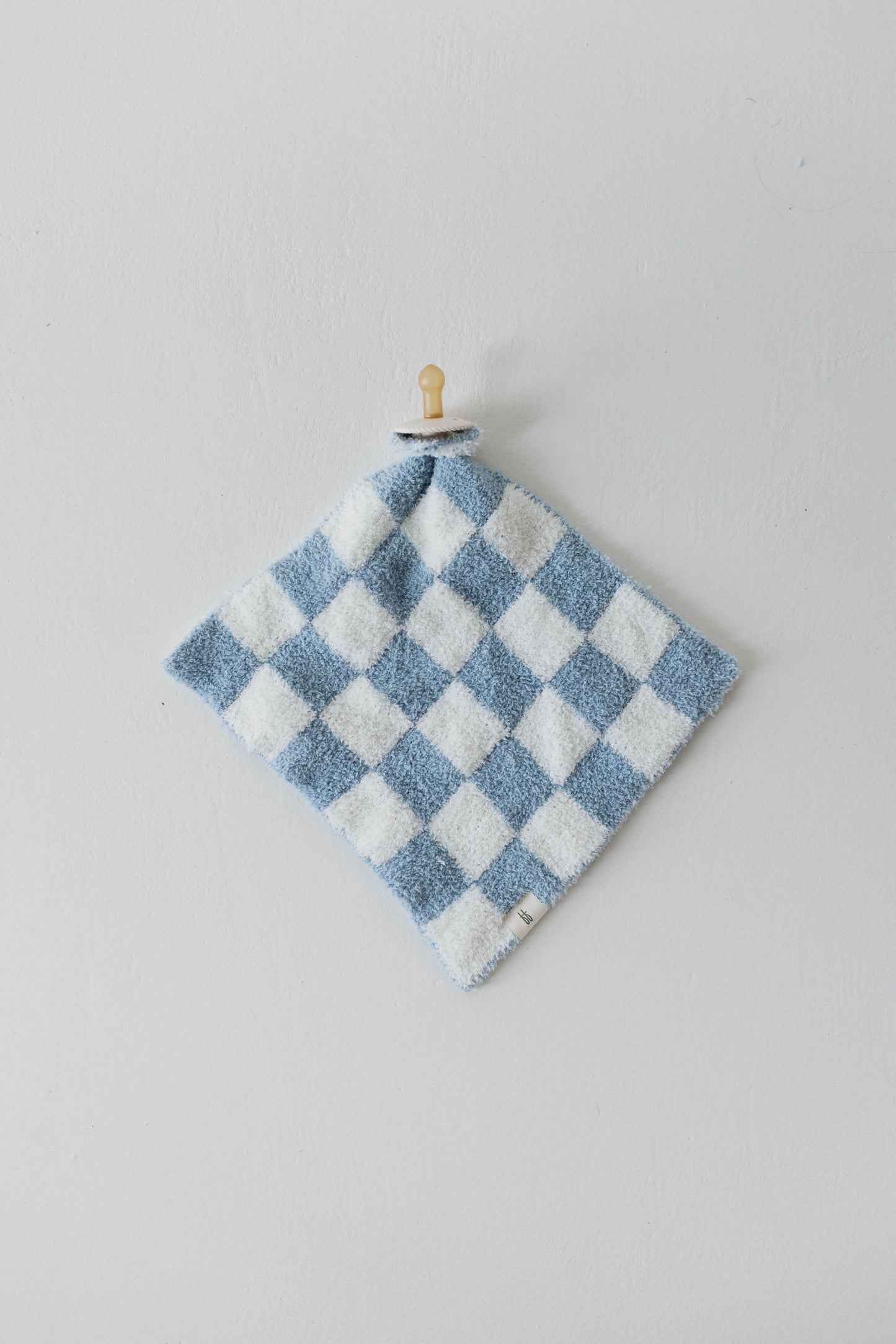 The Lovey | Sky Checkerboard by forever french baby, a blue and white checkered washcloth made from microfiber feather yarn with a soft, fluffy texture, is hanging from a small wooden peg on a light grey wall. The washcloth features a small fabric tag at the bottom corner, adding to its charming and practical design.