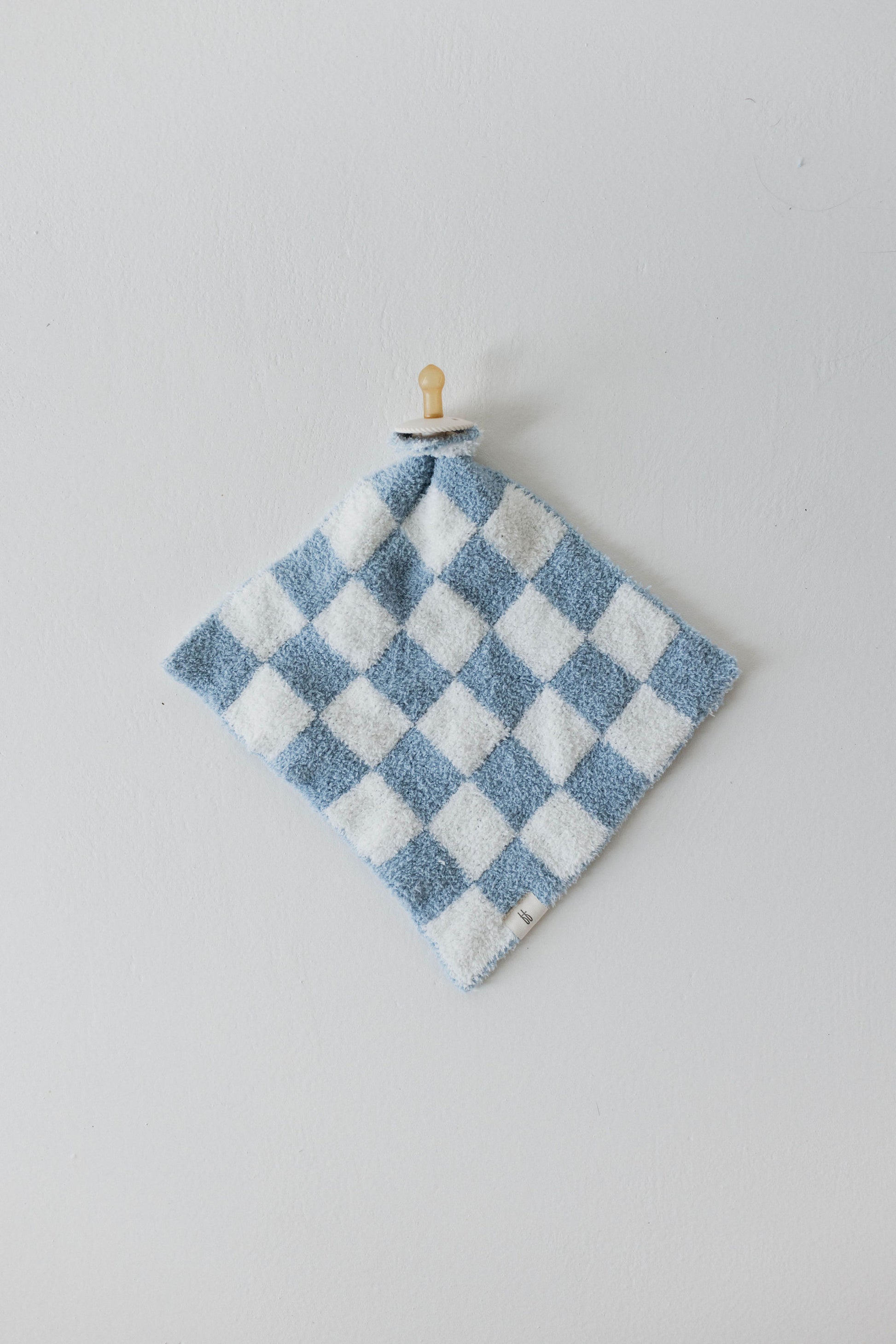 The Lovey | Sky Checkerboard by forever french baby, a blue and white checkered washcloth made from microfiber feather yarn with a soft, fluffy texture, is hanging from a small wooden peg on a light grey wall. The washcloth features a small fabric tag at the bottom corner, adding to its charming and practical design.
