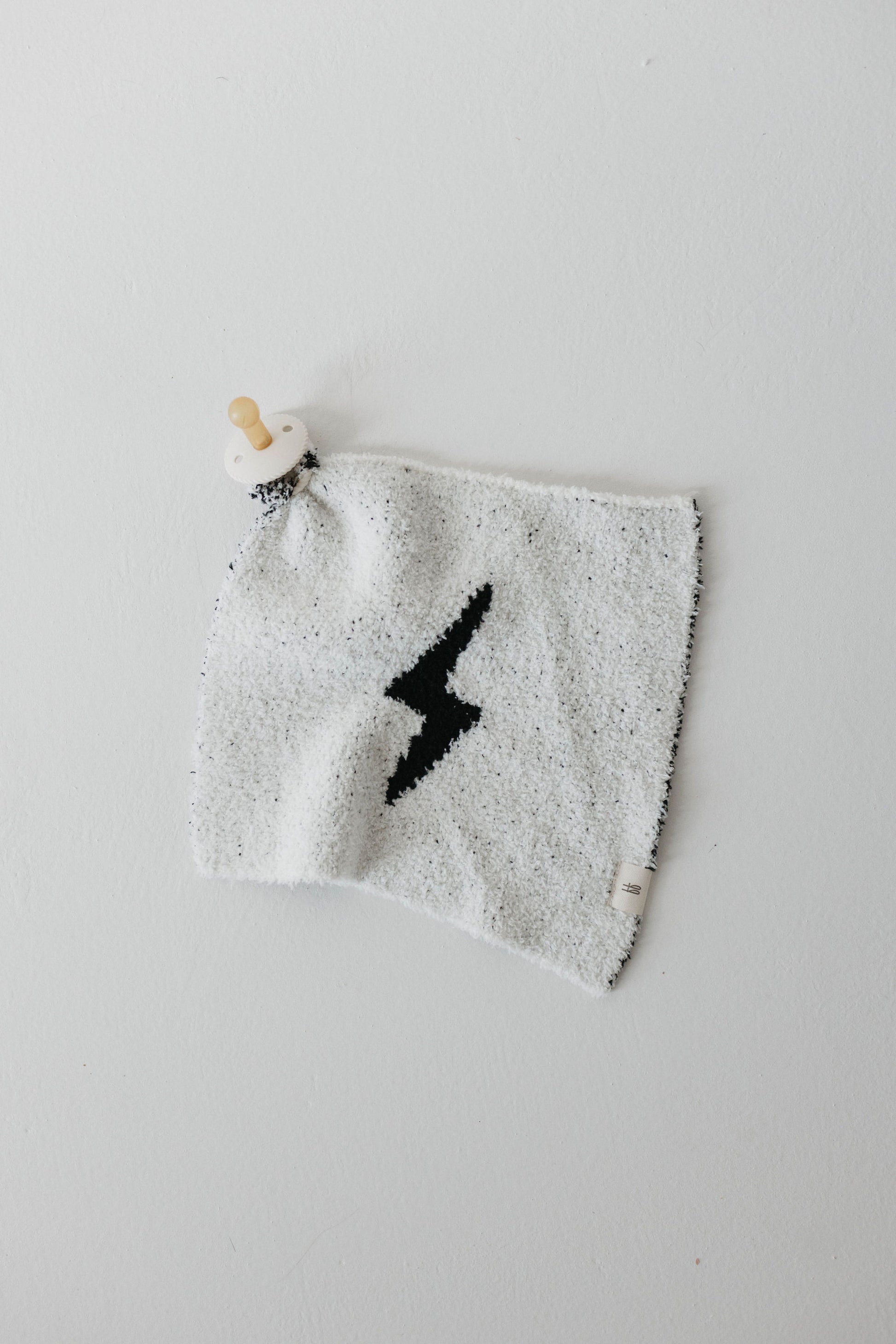 The "Lovey | Black & White Lightning Bolt" by Forever French Baby is a soft, white square blanket made from microfiber feather yarn. It features a black lightning bolt design in the center, with a wooden knob attached to one corner and a small tag sewn into one edge. This makes it perfect as a lovey for your little one.